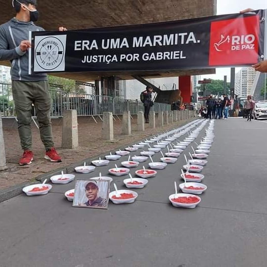 ato-era-só-uma-marmita-masp-marmitas-areia-vermelha pessoas segurando faixa escrito 'era uma marmita, justiça por gabriel' no vão do masp, em frente a 200 marmitas de isopor dispostas no chão