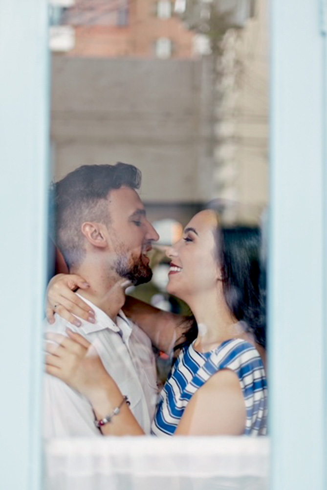 mayara e bruno sendo fotografados do lado de fora de uma janela. eles estão abraçados com os rostos quase colados um no outro e estão sorrindo
