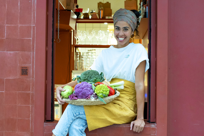 Bela Gil de camiseta branca, avental e calça jeans sentada na janela do Camélia Òdòdó segurando cesta de legumes.