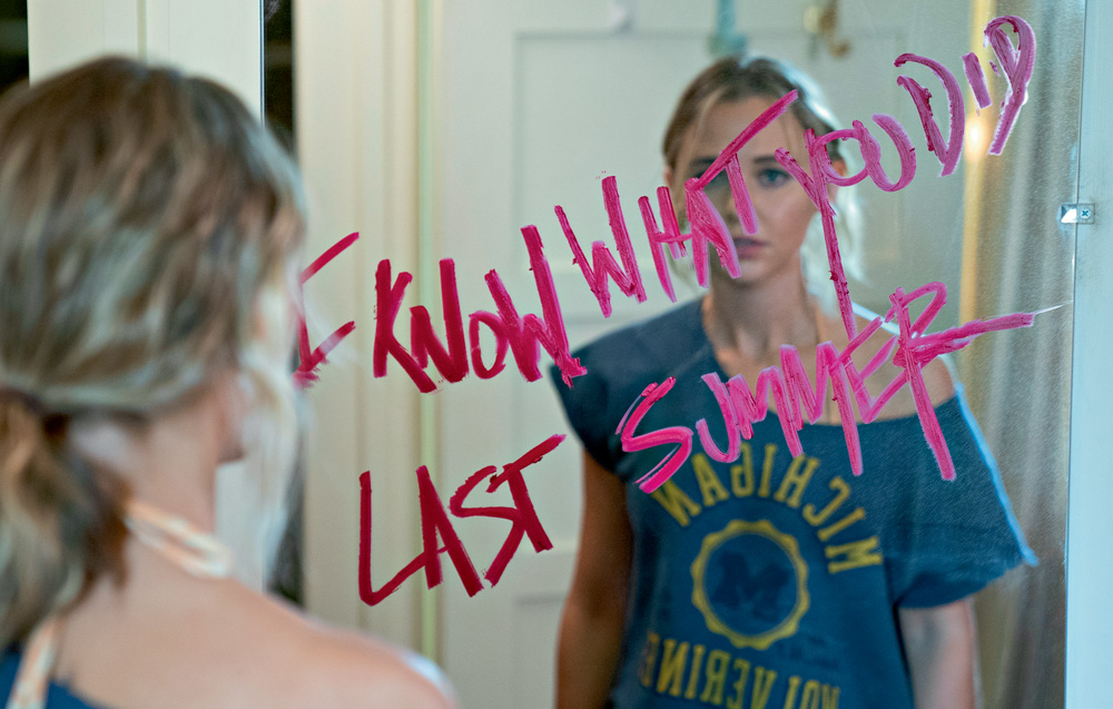 A imagem mostra uma jovem loira, em frente a um espelho, em que está escrito "Eu Sei o que Vocês Fizeram no Verão Passado" com batom.