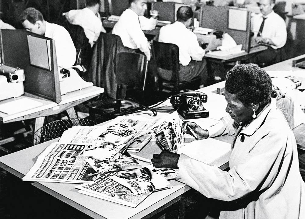 foto em preto e branco de carolina maria de jesus trabalhando na redação do jornal última hora, com outros colegas na sala. ela está lendo jornal