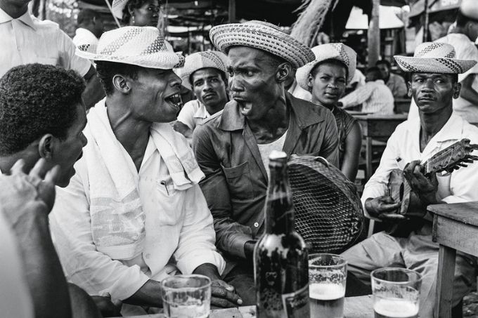 foto em preto e branco de grupo de homens de chapéu típico em bar, com cerveja nos copos da mesa, cantando e tocando instrumentos