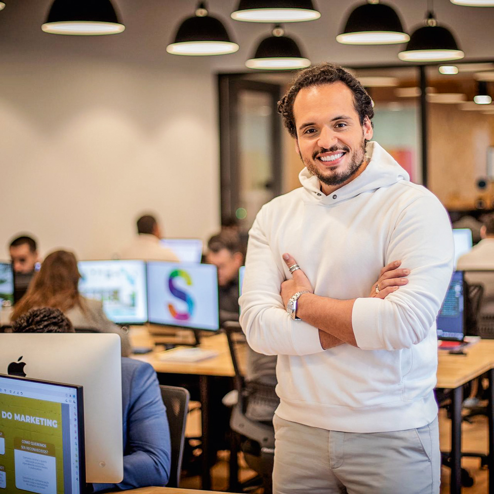 Bruno Sindona sorrindo de braços cruzados com computadores e pessoas trabalhando ao fundo