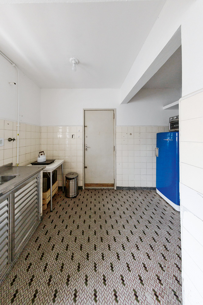 foto da cozinha de matheus ilt antes da reforma, com piso branco com detalhes pretos, parede branca e porta branca ao fundo e pia à esquerda