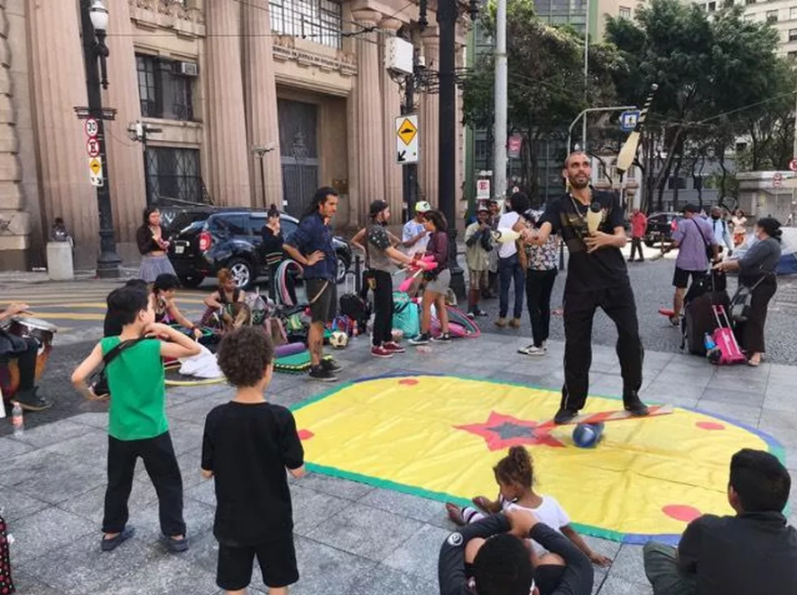Em uma praça, há crianças em volta de um circulo com um tapete colorido de brincar. Há um homem fazendo malabarismos no centro da roda