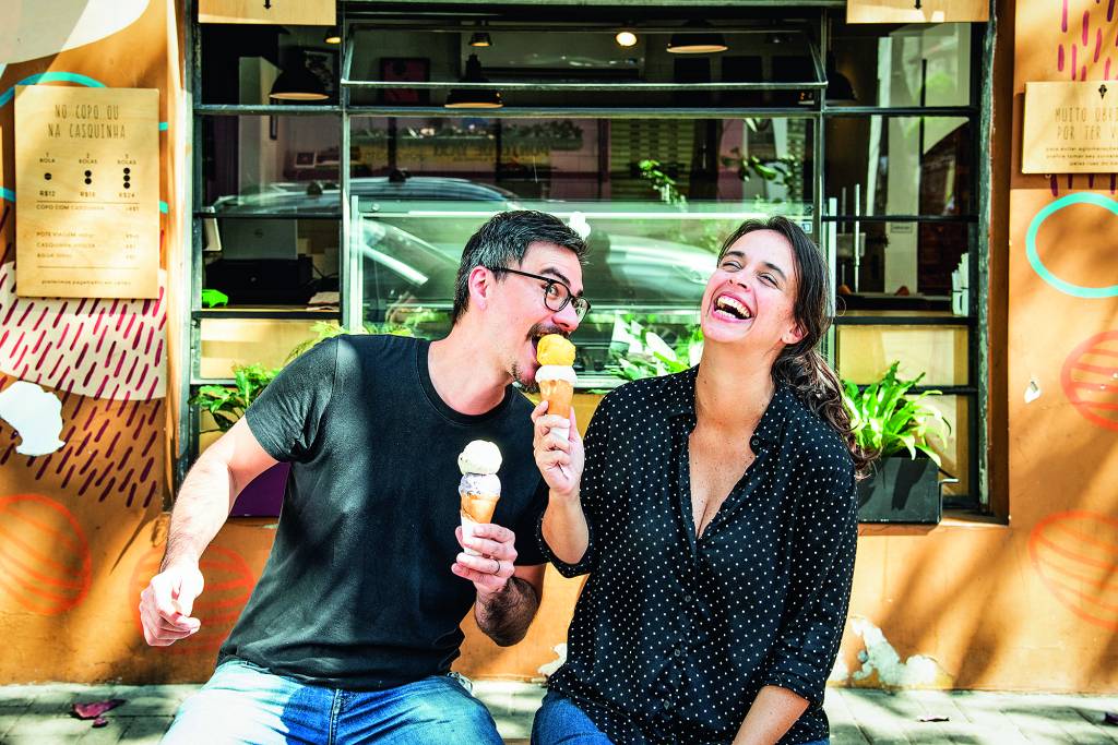 O casal Thomas Zander e Fernanda Bastos sentados em frente à Frida & Mina com ele tomando da casquinha de sorvete dela enquanto ela sorri.