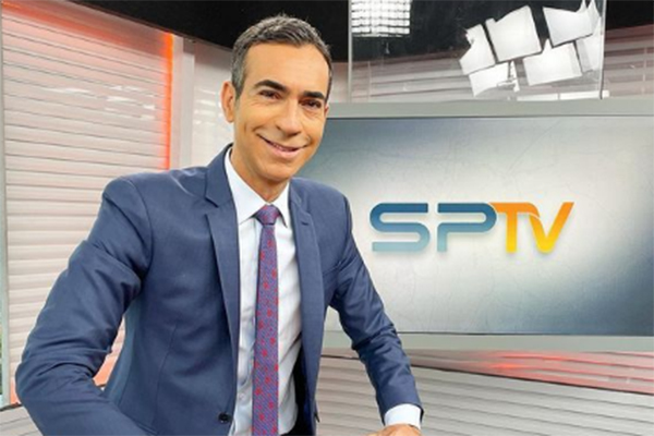 Imagem mostra Tralli sorrindo, de terno e gravata, na frente de televisão, onde pode-se ler: SPTV