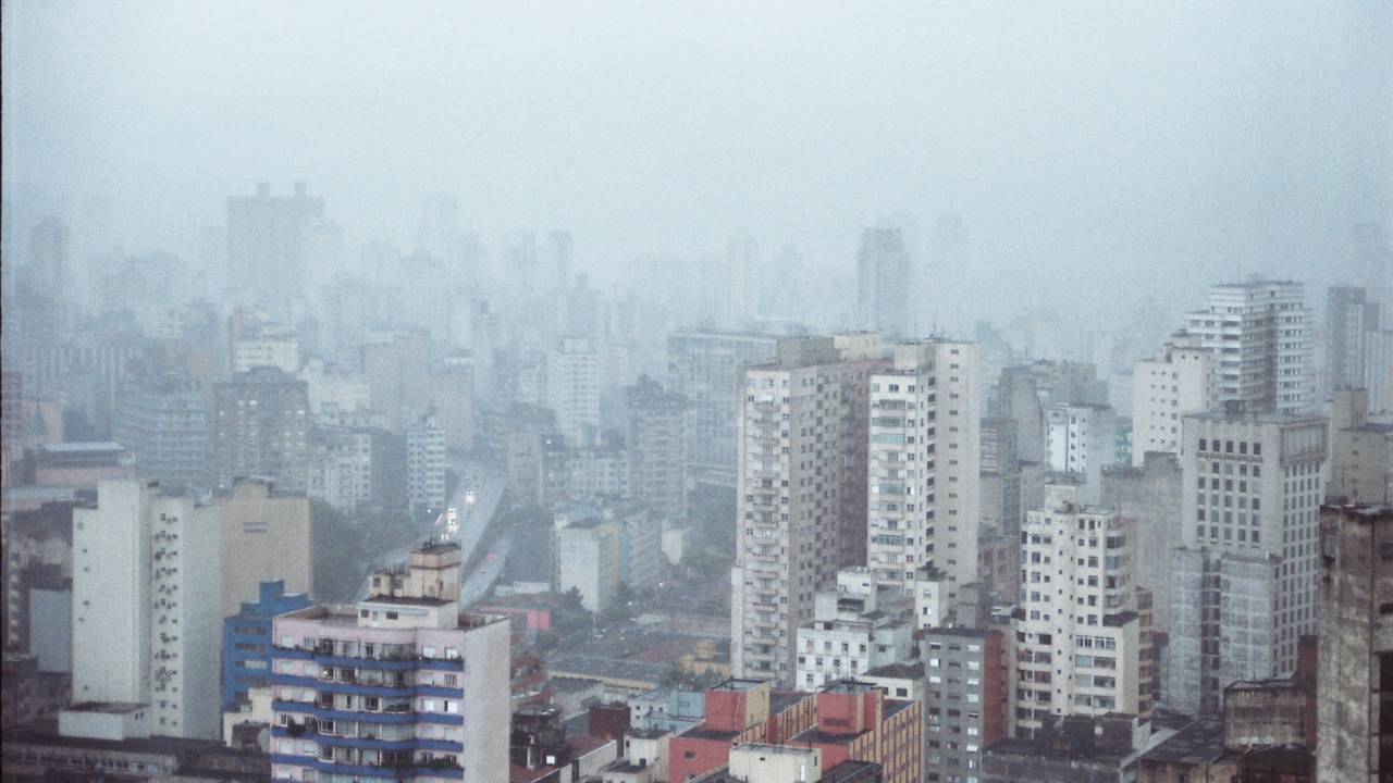Imagem mostra horizonte de prédios encobertos com tempo nublado e chuva ao longe.
