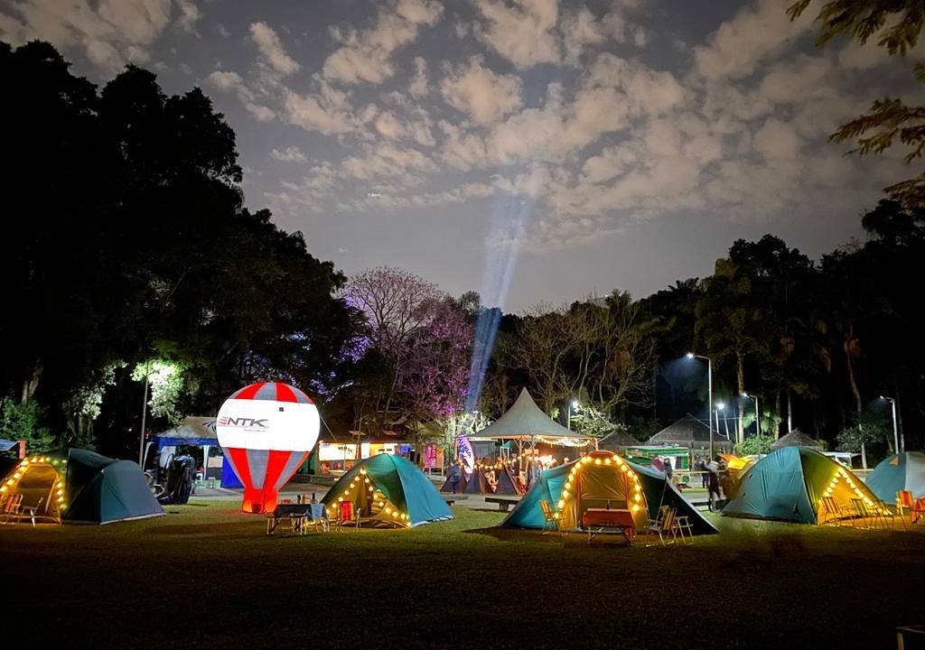 À noite, barracas com luzes de LED penduradas são dispostas em um parque. Dá para ver coqueiros e o céu ao fundo. Também há uma luz em direção ao céu