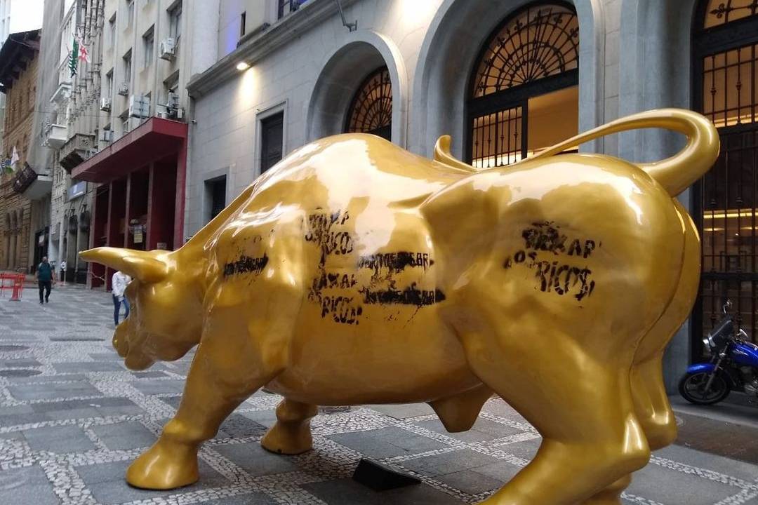 A imagem mostra o touro dourado, uma escultura no centro de São Paulo, com pichação escrita #taxar os ricos"