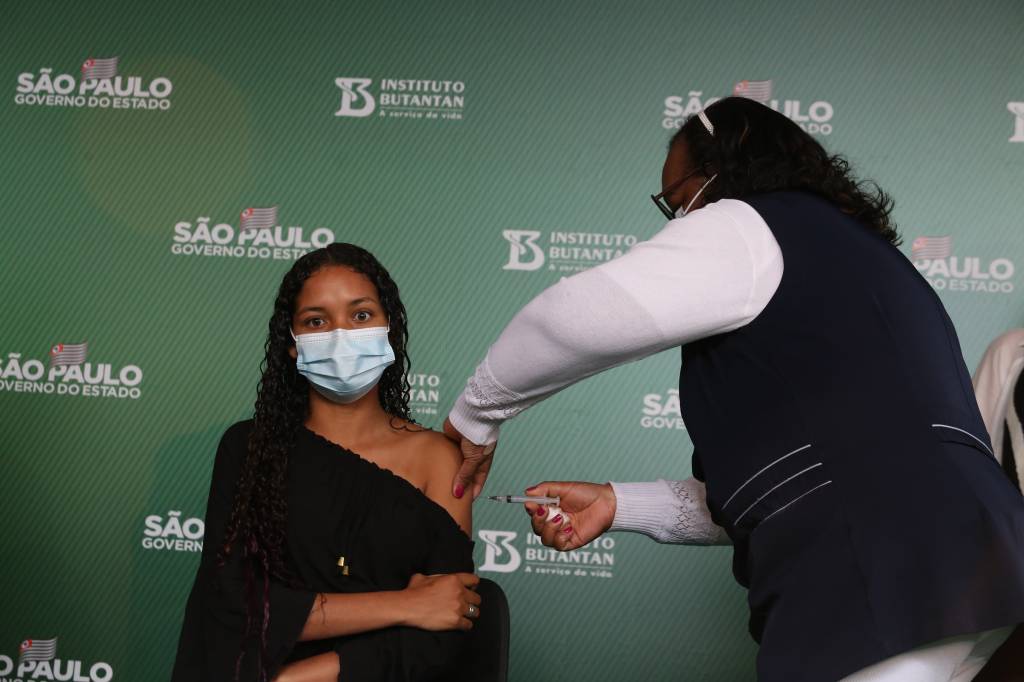 Uma enfermeira vacinando uma menina. Ao fundo, um painel verde com o logo do Governo do Estado e do Instituto Butantã.