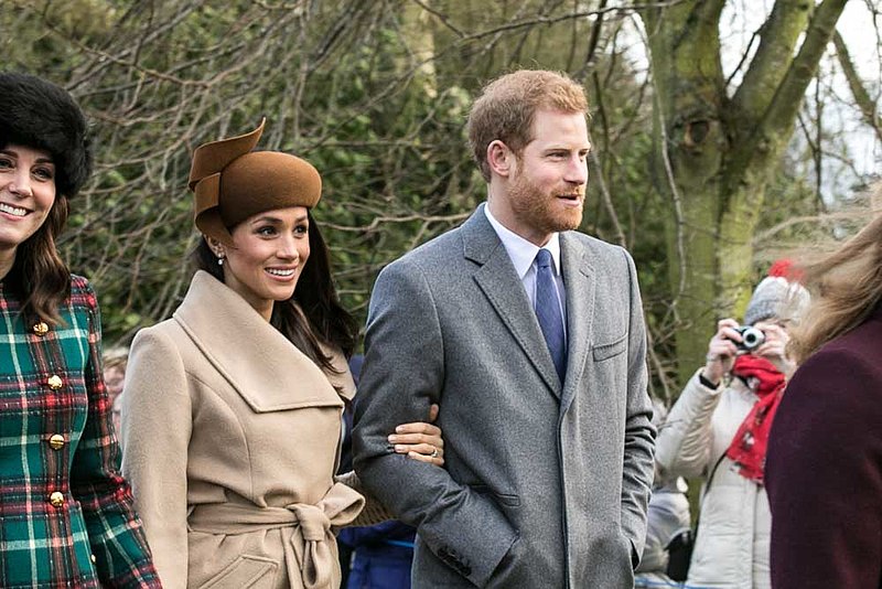Harry e Meghan estão um ao lado do outro e ela segura seu braço. Meghan veste um casaco de inverno e um chapéu marrons e Harry usa um terno cinza e uma gravata azul.