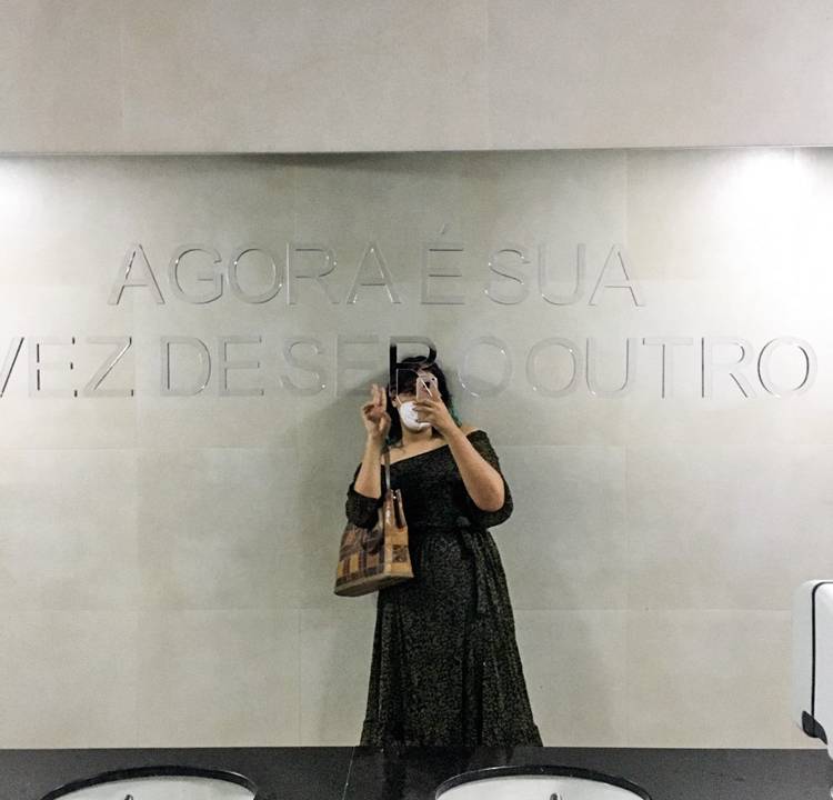 Imagem tirada em espelho de banheiro com os dizeres (no espelho): "Agora é sua vez de ser o outro". A pessoa que tira foto aparece no centro do reflexo, de vestido preto.