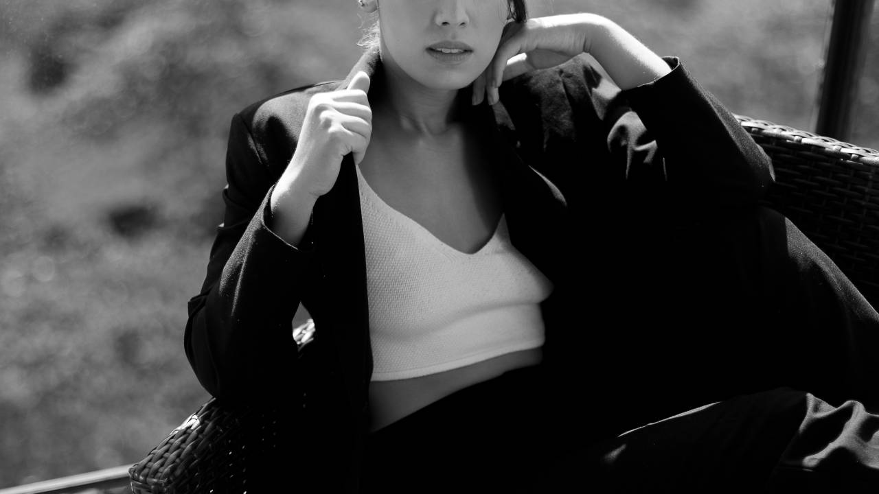 Ana Hikari em foto preto e branco aparece sentada em frente a vidraça e veste paletó com blusa.