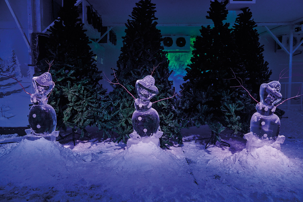 Em um espaço temático iluminado com luz negra, a neve no chão e árvores de pinheiro atrás. No centro da imagem, esculturas de gelo do personagem Olaf, de Frozen