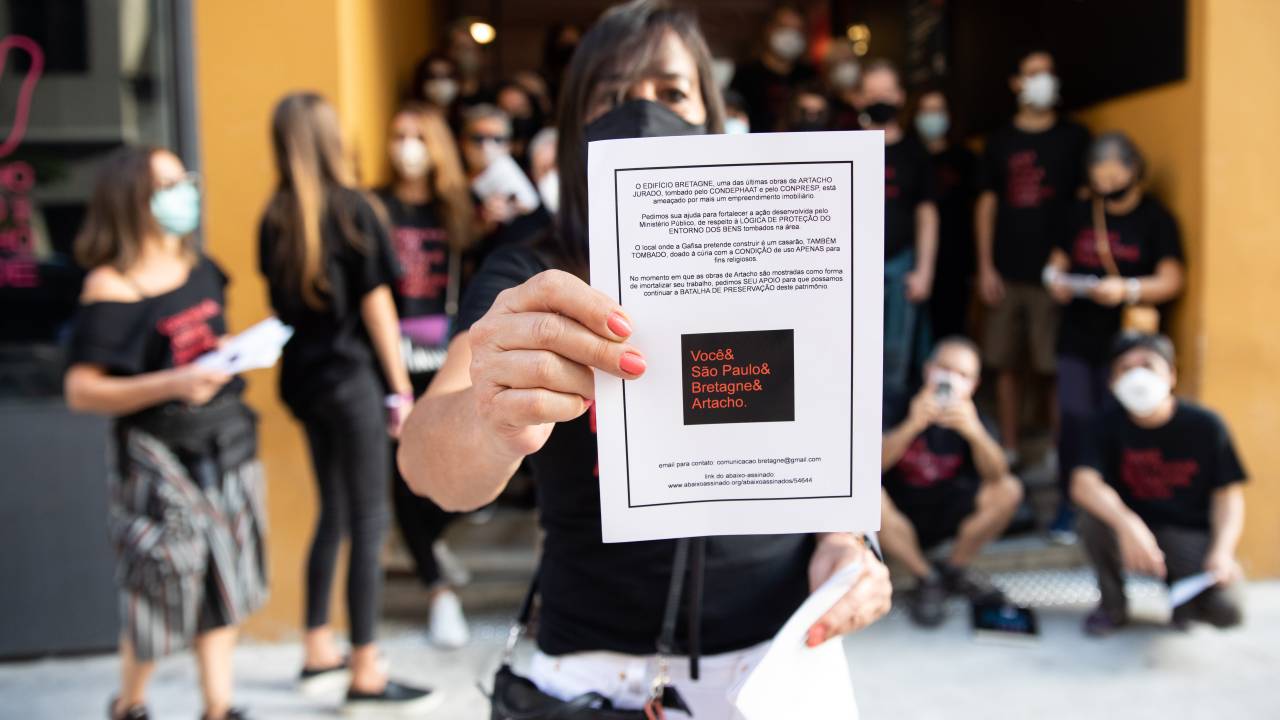 Imagem mostra uma mulher segurando uma carta, em primeiro plano. Atrás dela, aparece um grupo de cerca de trinta pessoas, todas vestindo camisetas pretas