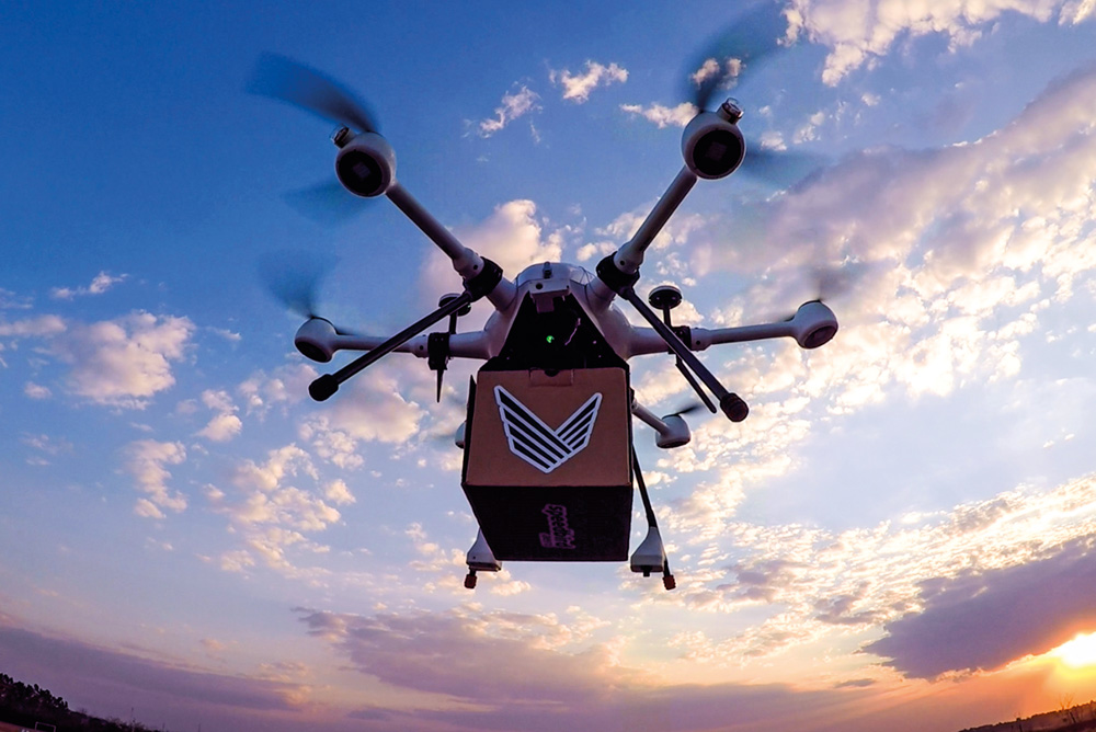 A imagem mostra um drone no céu com uma caixinha em baixo preso nele.