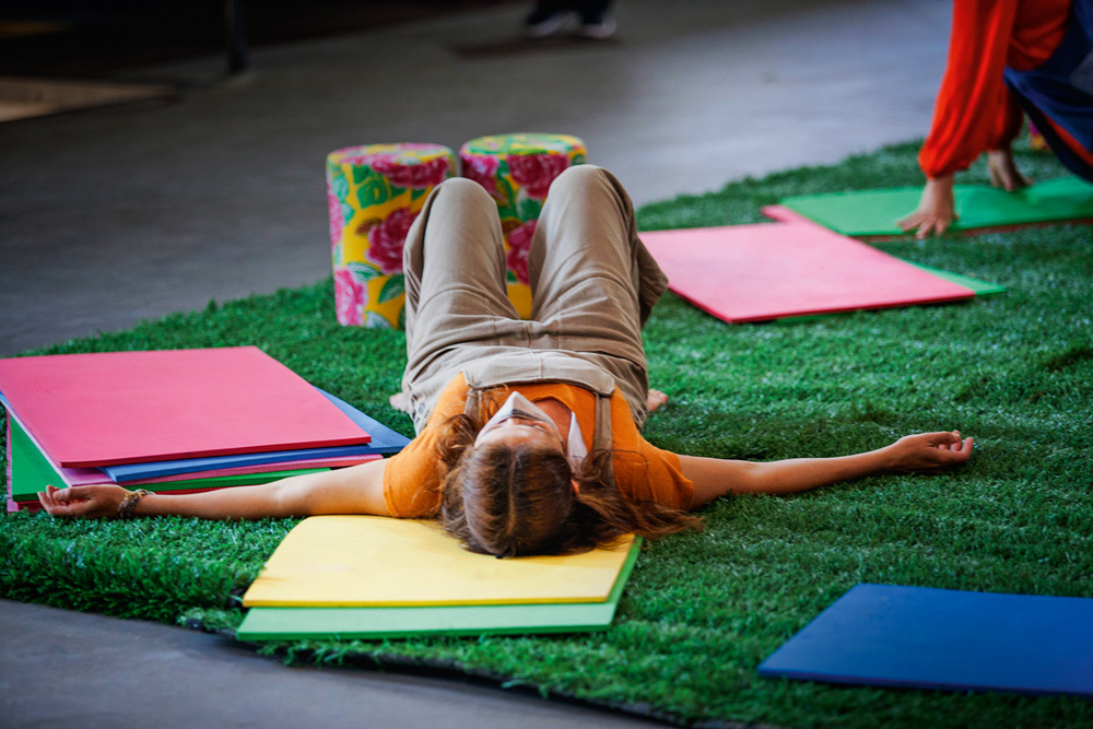 Em um tapete de grama artificial, uma mulher deita, de costas para a câmera, com várias cartolinas coloridas ao seu redor