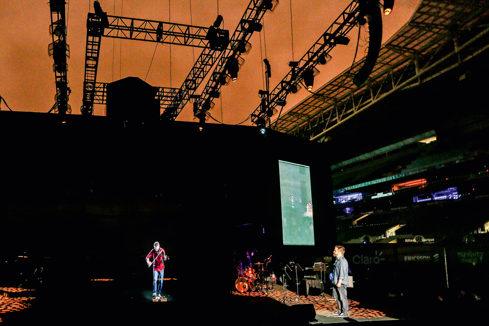 A imagem mostra um palco e um holograma de Lucas Lima em cima dele. O palco está escuro mas é possível ver claramente o cantor.