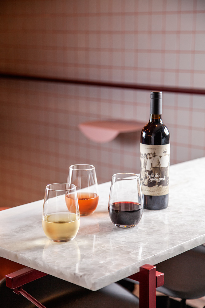 Sobre uma bancada de mármore, há uma garrafa de vinho tinto, junto de três copos de vidro, um com vinho rosé, um com vinho branco e outro, com tinto