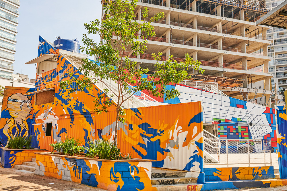 Imagem mostra casa com parede toda grafitada das cores amarelo, laranja e azul. Ao fundo, um prédio em construção.