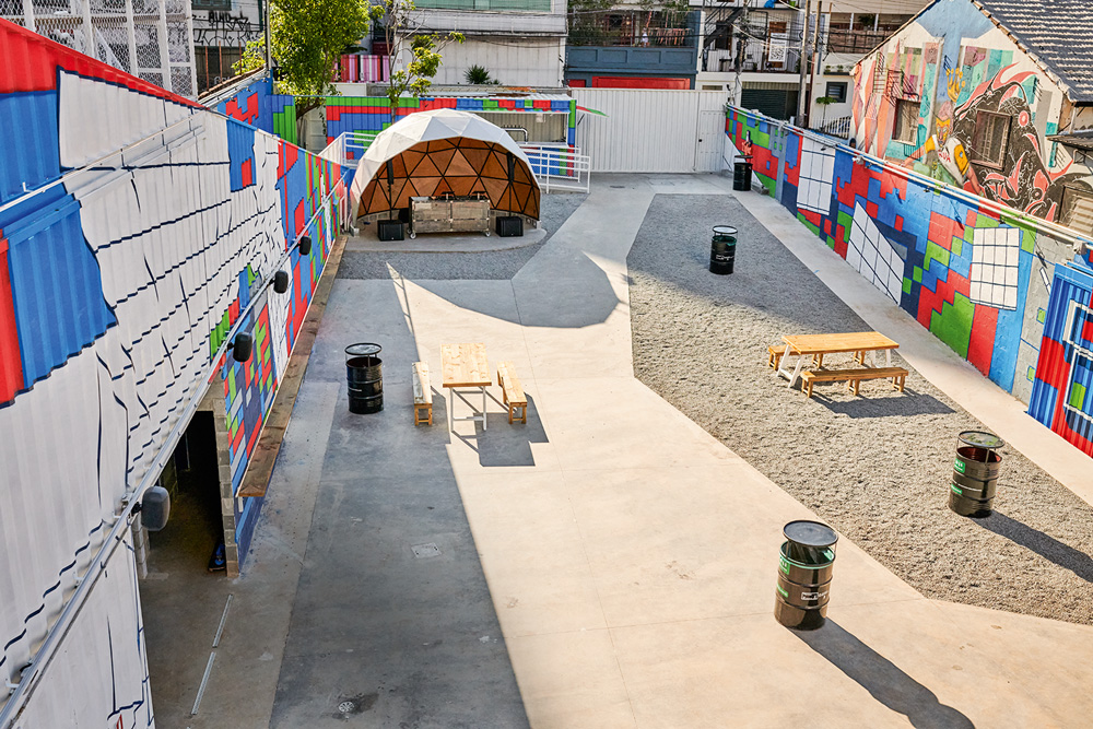 Imagem mostra praça de concreto com mesas, barris pretos, uma cúpula geodésica e paredes com cubos coloridos desenhados.