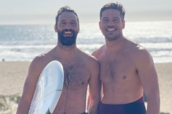 Imagem mostra dois homens, um ao lado do outro, em uma praia, o da esquerda segurando uma prancha de surfe.