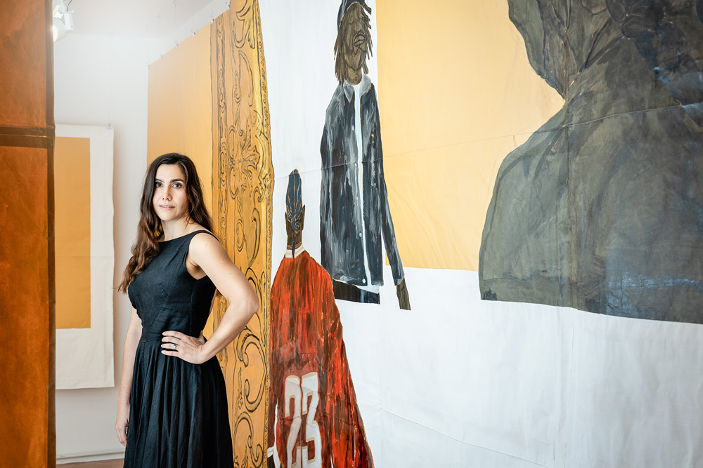 Uma mulher de vestido preto posa com a mão na cintura em frente a uma parede que possui uma obra de arte nela