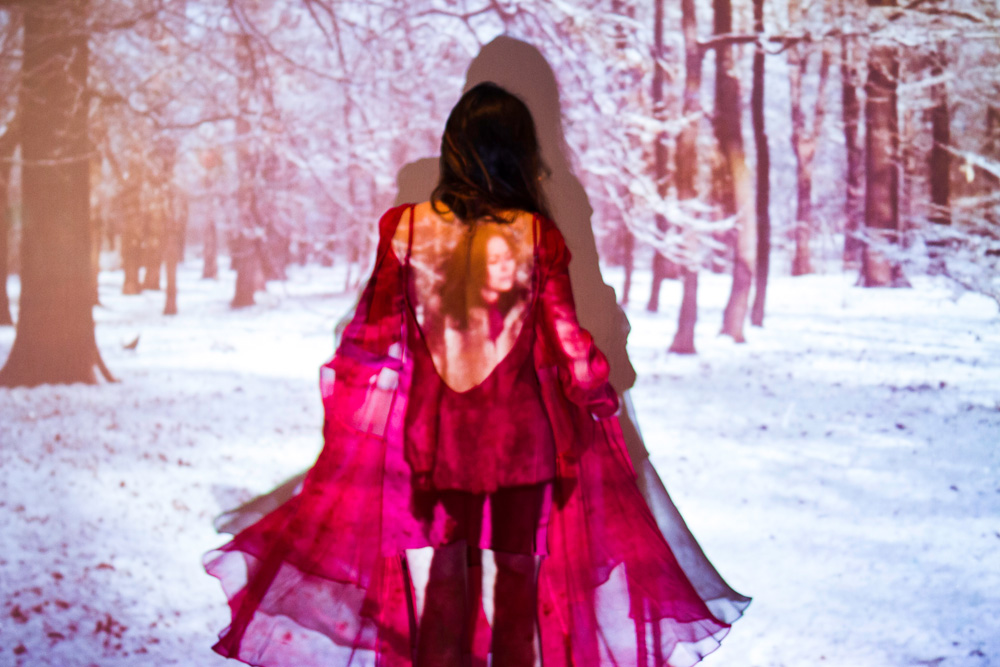 Em um palco de teatro, há uma projeção de um cenário com árvores e muita neve. Uma mulher está no centro da foto de costas com os braços abertos e um vestido rosa