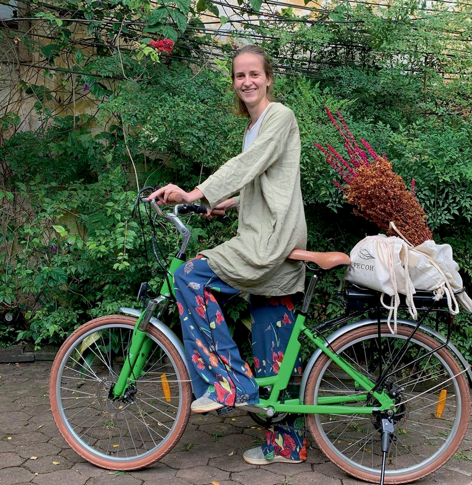A imagem mostra Elise em cima de uma bicicleta, apoiada com um pé. Ela sorri para a câmera em um ambiente cheio de plantas.