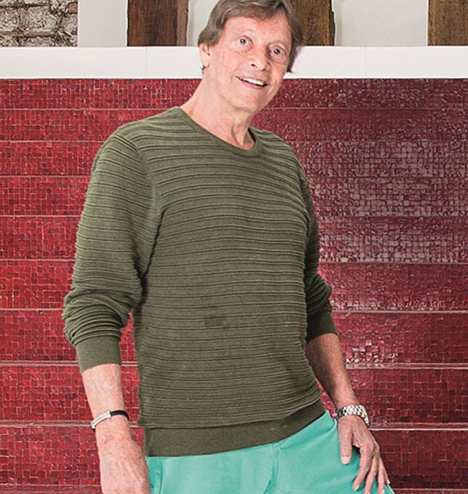 Fabio Porchat posa de pé sorrindo. Veste suéter verde-escuro e calça verde-água.