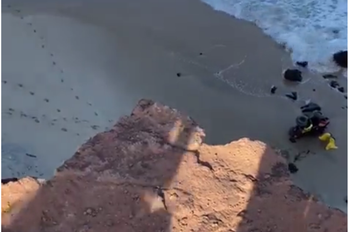 A imagem mostra a visão de cima da falésia, podendo ver a praia e o quadriciclo deitado na areia.