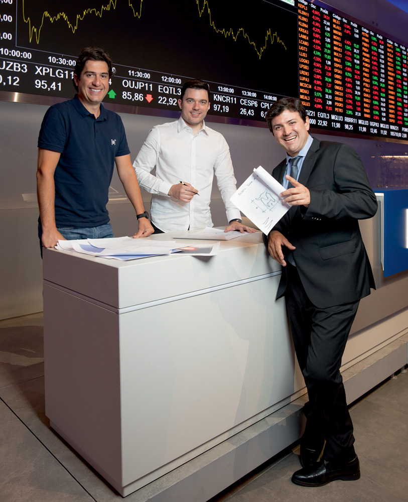 A partir da esquerda, Felipe, Rafael e Pablo sorriem reunidos em um balcão branco. Ao fundo, painéis da bolsa de valores.