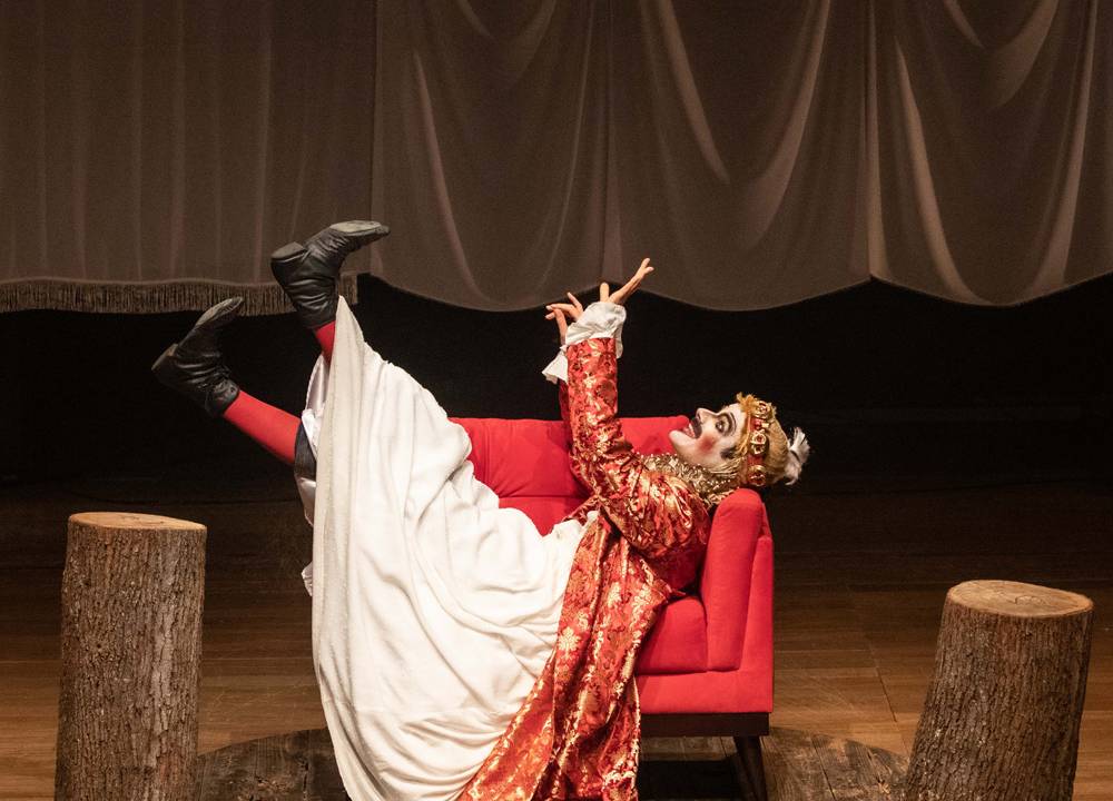 Em um palco de teatro, homem vestido e maquiado de rei Henrique IV está deitado em um sofá com as pernas e mãos para o ar, articulando