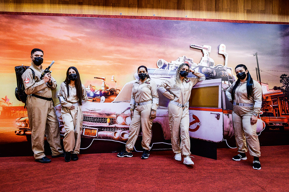 Em frente a um painel que mostra cenário do filme Ghostbusters, cinco pessoas vestidas de caça-fantasmas posam para a foto