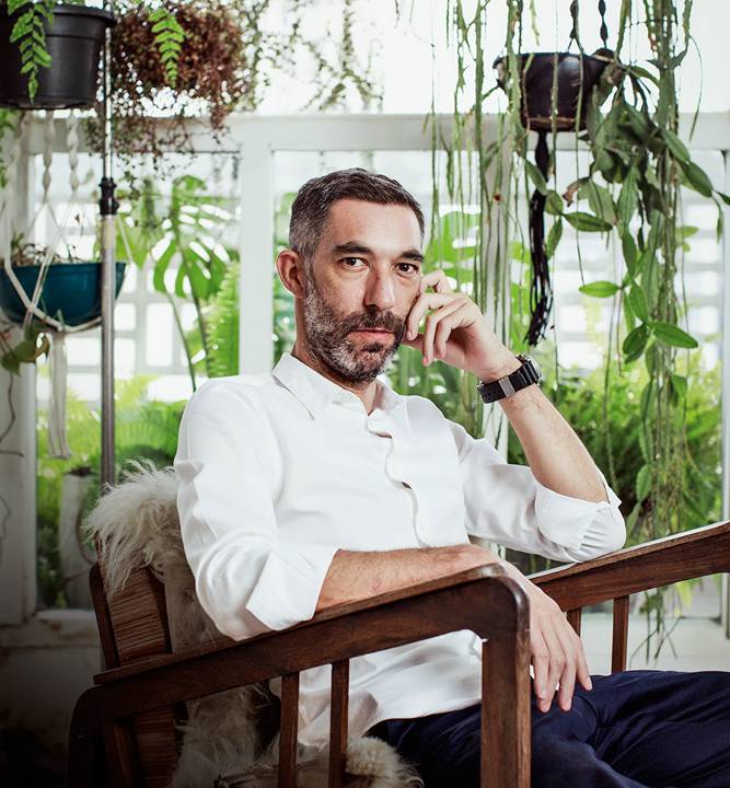 Greg Bousquet posa vestindo camisa branca e sentado em cadeira de madeira. Uma das mãos está apoiada no rosto. Ao fundo, plantas penduradas em janelas de vidro.