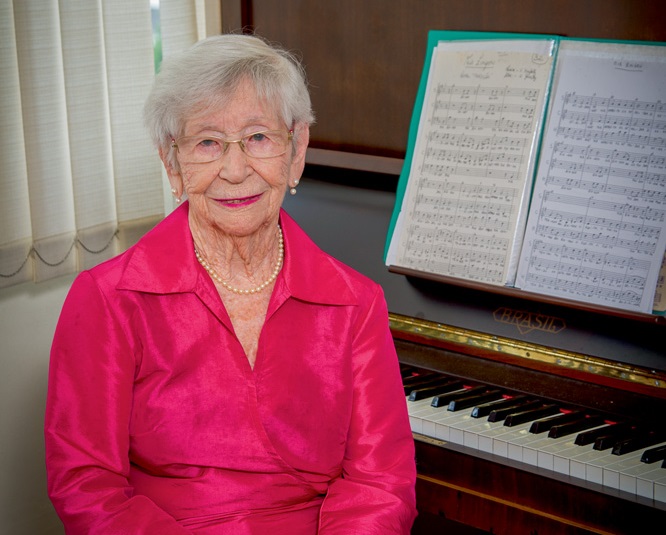 Uma senhora grisalha de 95 anos posa sentada, com camisa social rosa, em frente a um piano. Há uma partitura aberta no instrumento