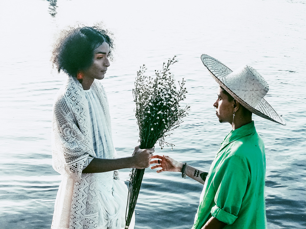Imagem mostra duas pessoas, uma de vestido branco e outra de cmaisa verde, se olhando enquanto uma entrega flores à outra. No fundo, o mar.