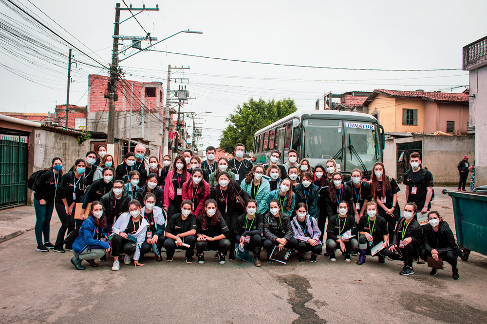 Um grupo grande de alunos posam enfileirados para a câmera em uma comunidade. Estão todos de máscaras contra a Covid