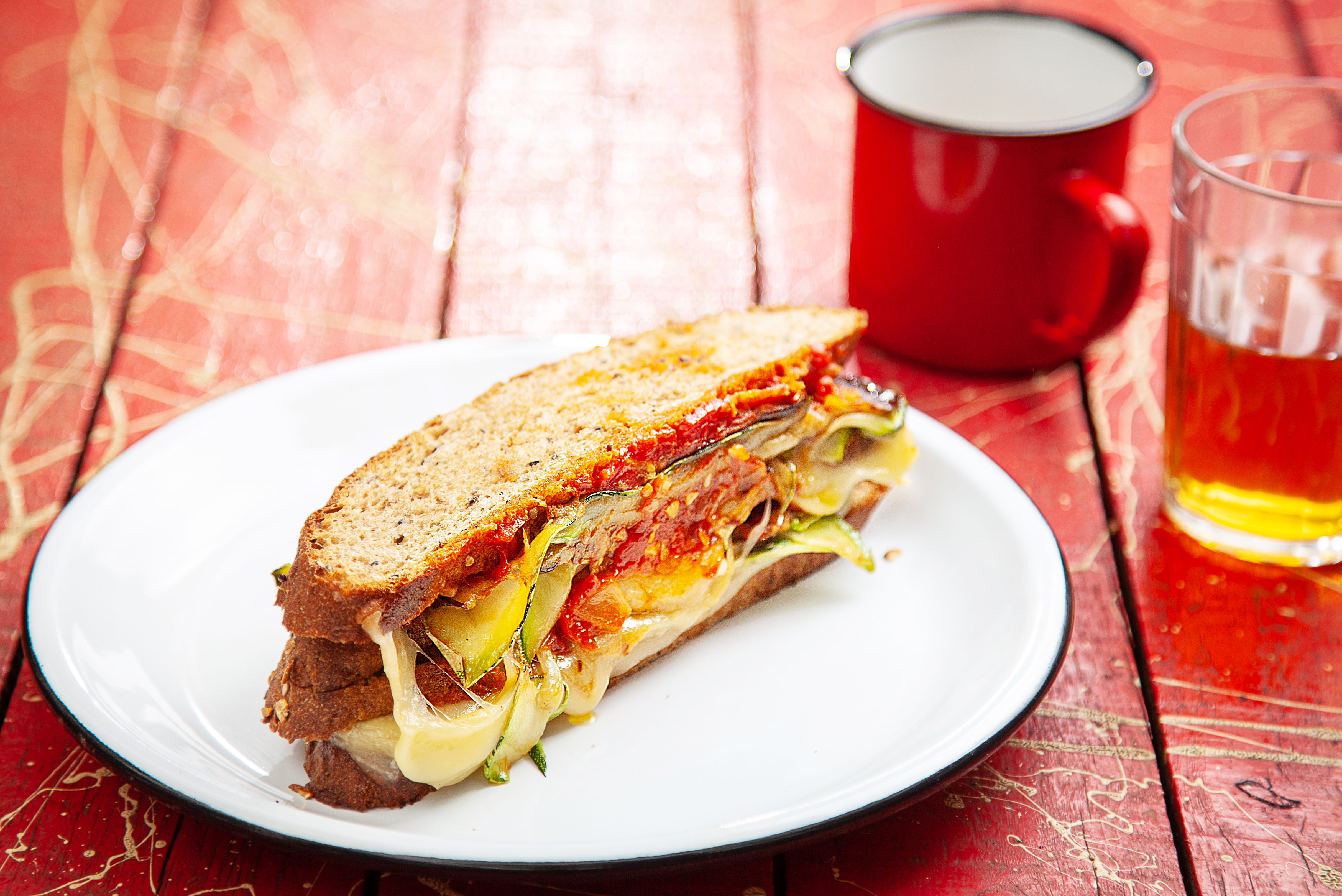 Sanduíche com queijo e molho de tomate sobre prato branco em cima de mesa de madeira pintada de vermelho.