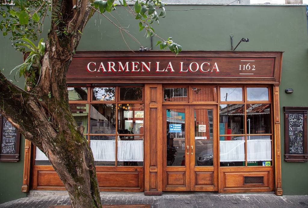 Fachada do restaurante Carmen La Loca, com porta de madeira, parede verde musgo e árvore na lateral esquerda.