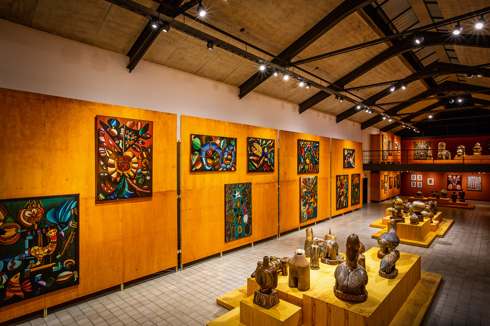 Imagem mostra sala com painel de pinturas e esculturas expostas.