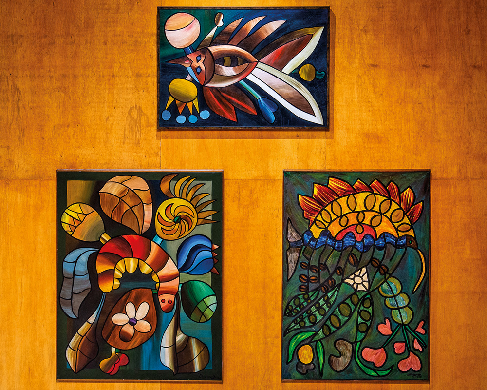 Imagem mostra três pinturas expostas em parede. As três são coloridas e mostram animais e plantas.