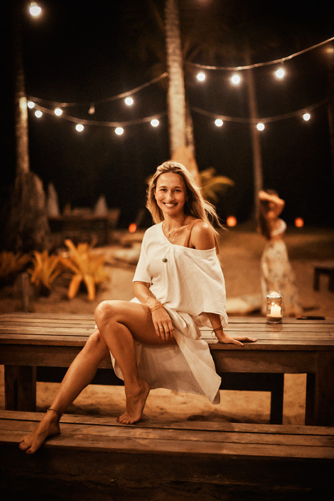 A imagem mostra Joice, de vestido branco, sentado em banco de madeira.