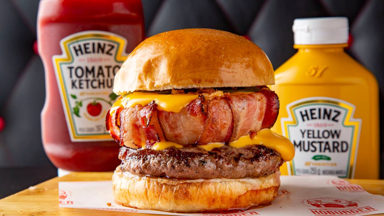 Hambúrguer com camada alta de bacon em frente à vidros de ketchup e mostarda da marca Heinz.
