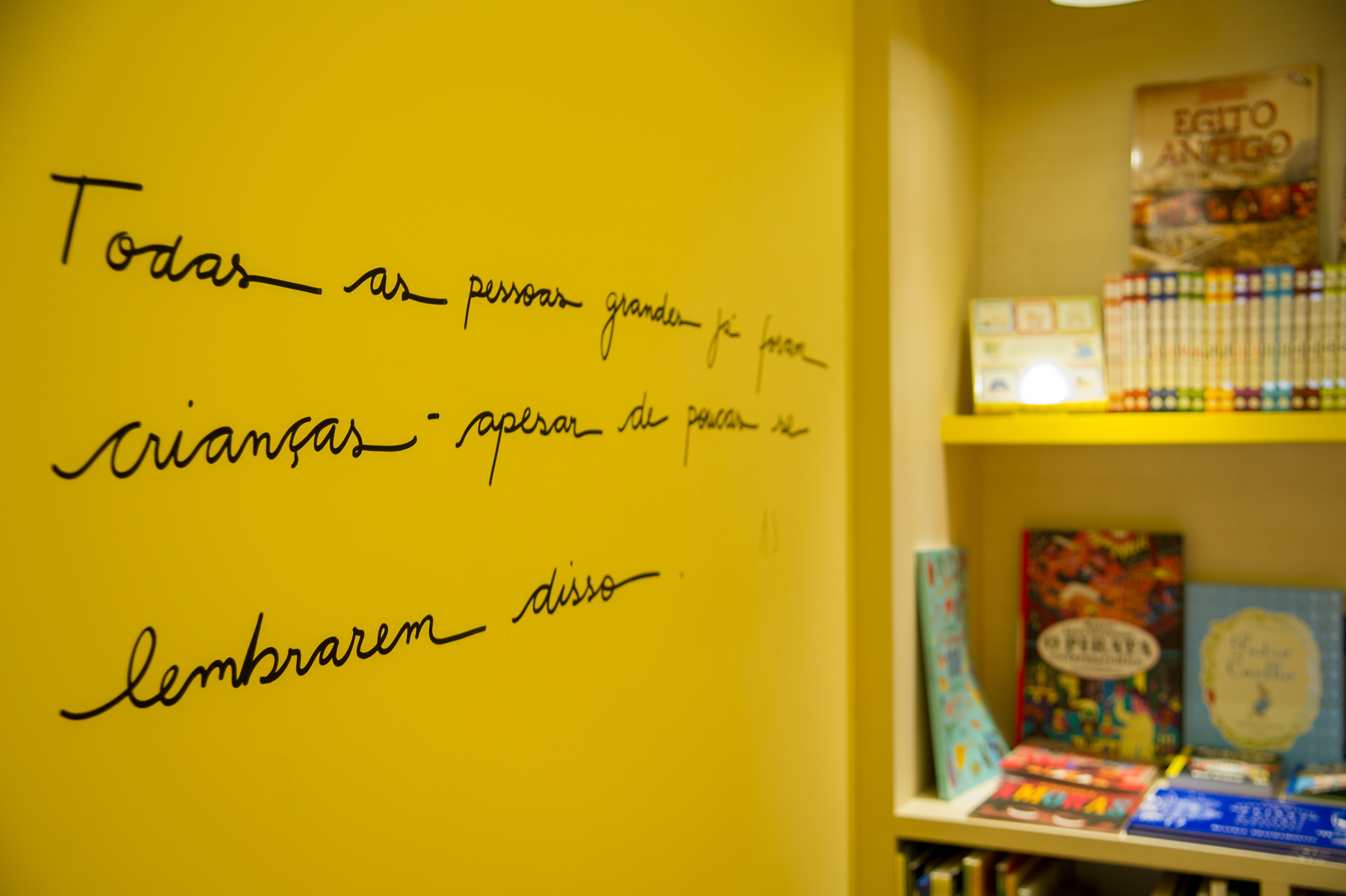 Foto em detalhe exibe frase escrita em uma parede branca. À direita, alguns livros em prateleiras.
