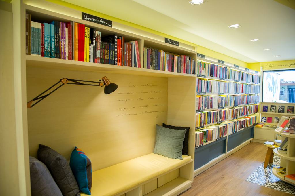 Livraria do Brooklin (Rogério Pallatta) Sofázinho embutido entre prateleiras de livros tem luz e almofadas. Móveis na cor de madeira clara.