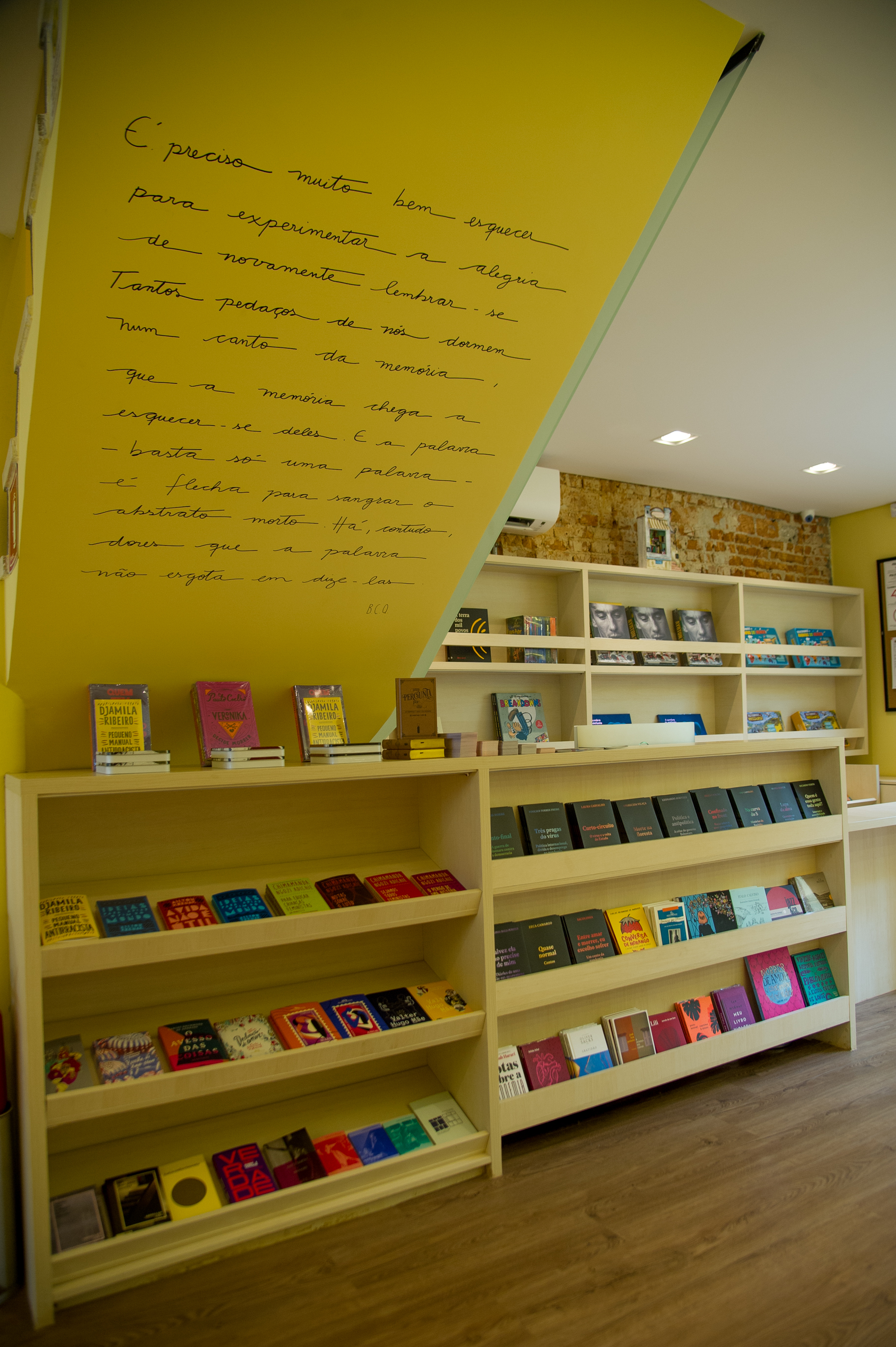 Foto exibe teto na diagonal com grande texto escrito na parede branca. Abaixo, grande prateleira de madeira com livros.