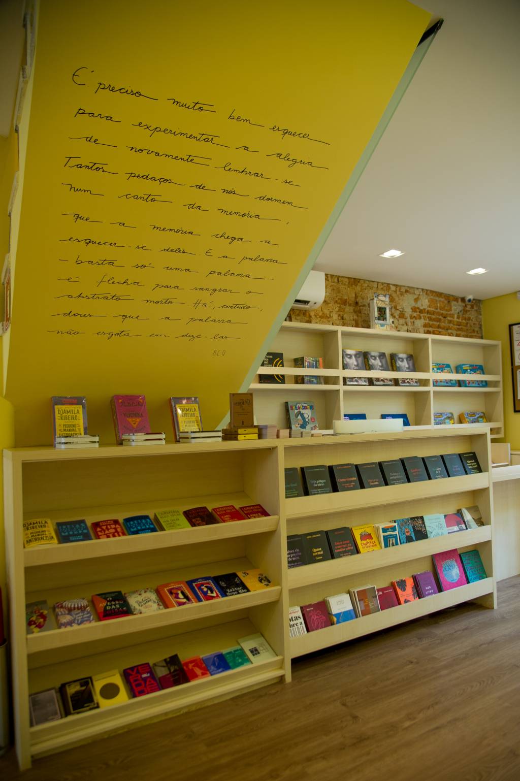 Livraria do Brooklin (Rogério Pallatta) Foto exibe teto na diagonal com grande texto escrito na parede branca. Abaixo, grande prateleira de madeira com livros.