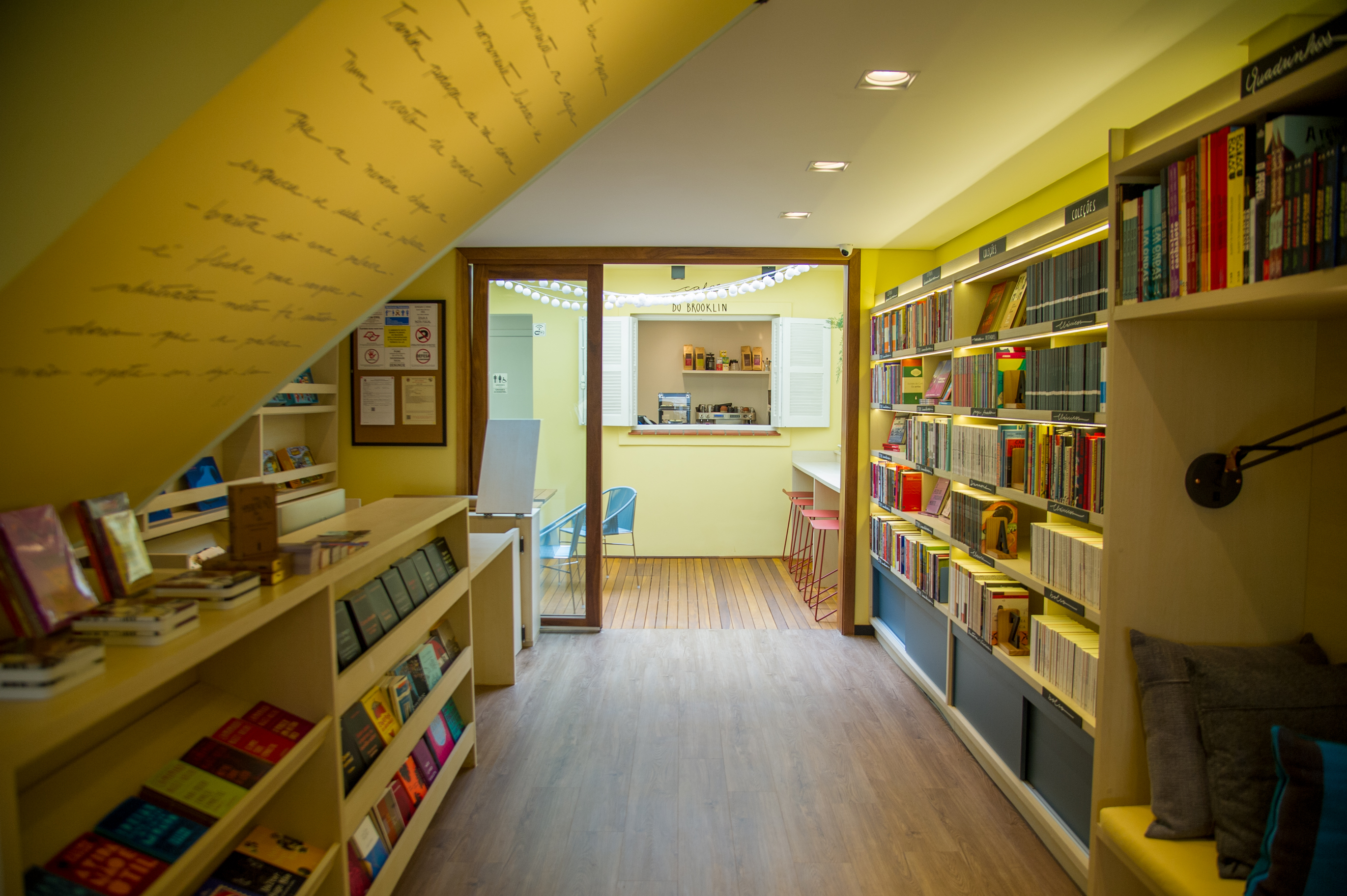 Foto exibe de modo centralizado o espaço da livraria, com chão de madeira, luzes embutidas no teto e duas prateleiras nas duas paredes.